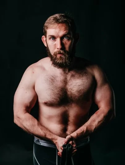 A bearded, shirtless man with chest hair standing against a dark background
