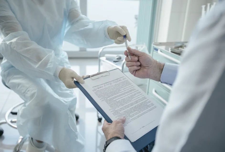 A physician holding a clipboard as a patient in a gown signs a form