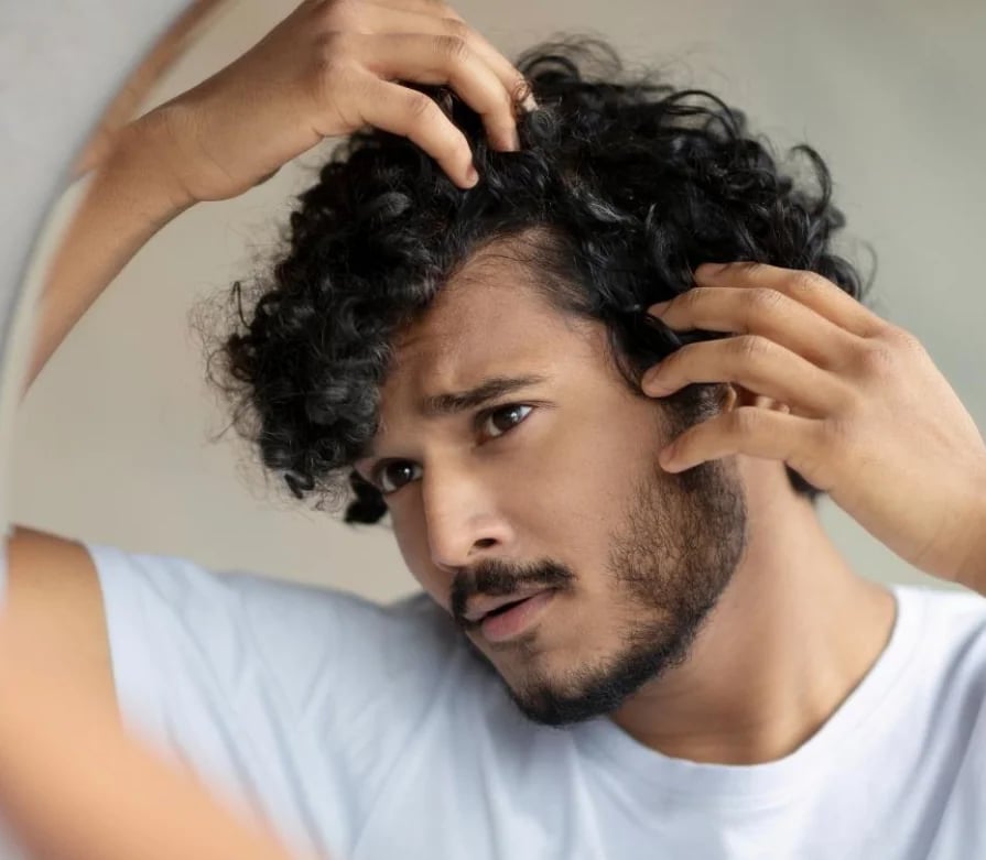A man with curly dark hair checking his hairline in a mirror