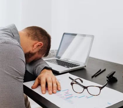 Tired man resting his head on his arm at a desk beside a laptop and a pair of glasses